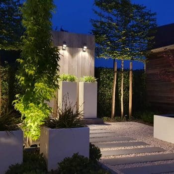Indirecte tuinverlichting verlicht de verschillende plantbakken en pad met stapstenen dat uitgeeft op een houten scherm in padoek met drie hoge plantbakken als blikvanger in deze moderne en gezellige tuin.